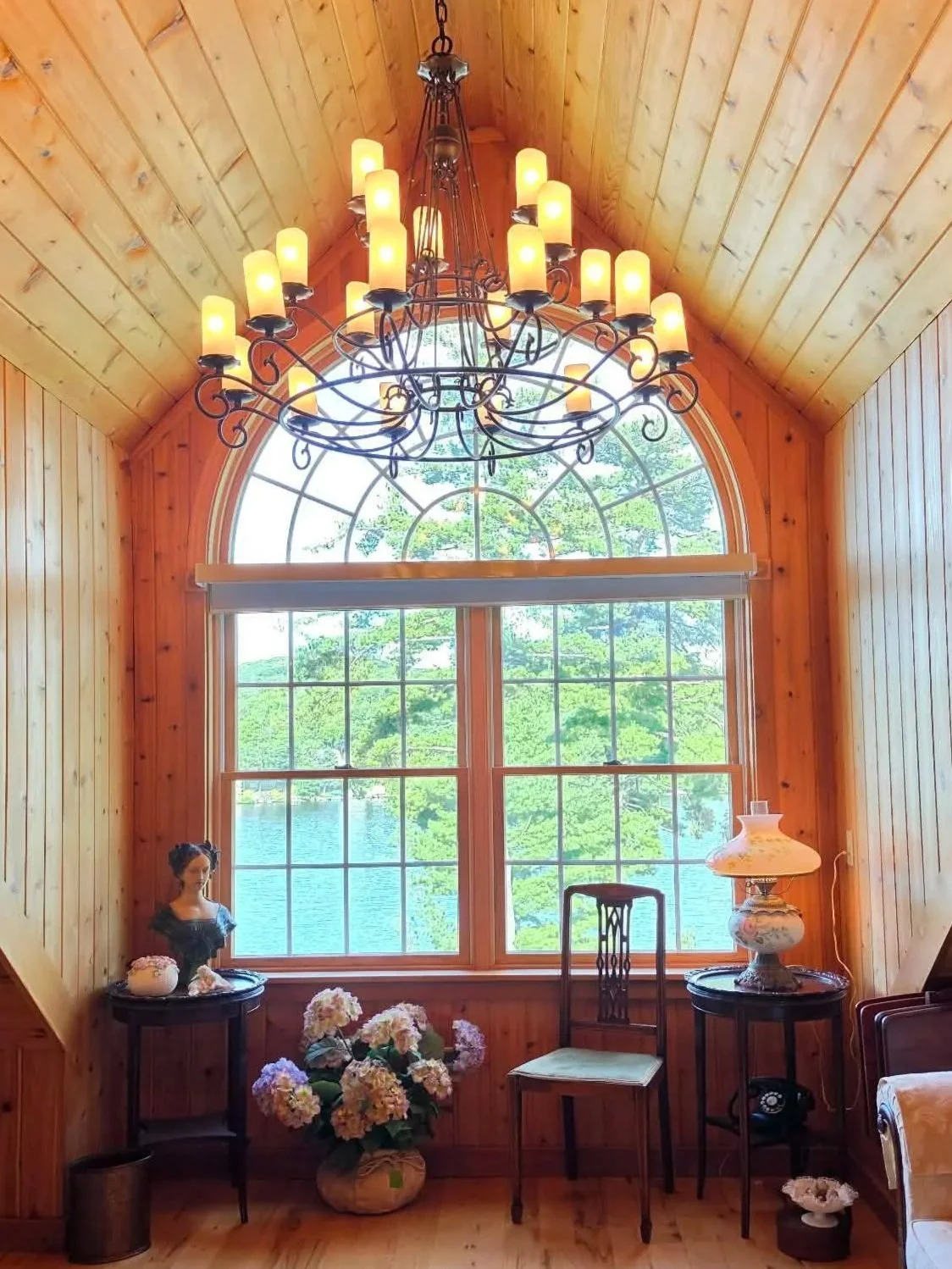 Wood-paneled estate interior with antique furnishings, a chandelier, and an arched window overlooking a lake.