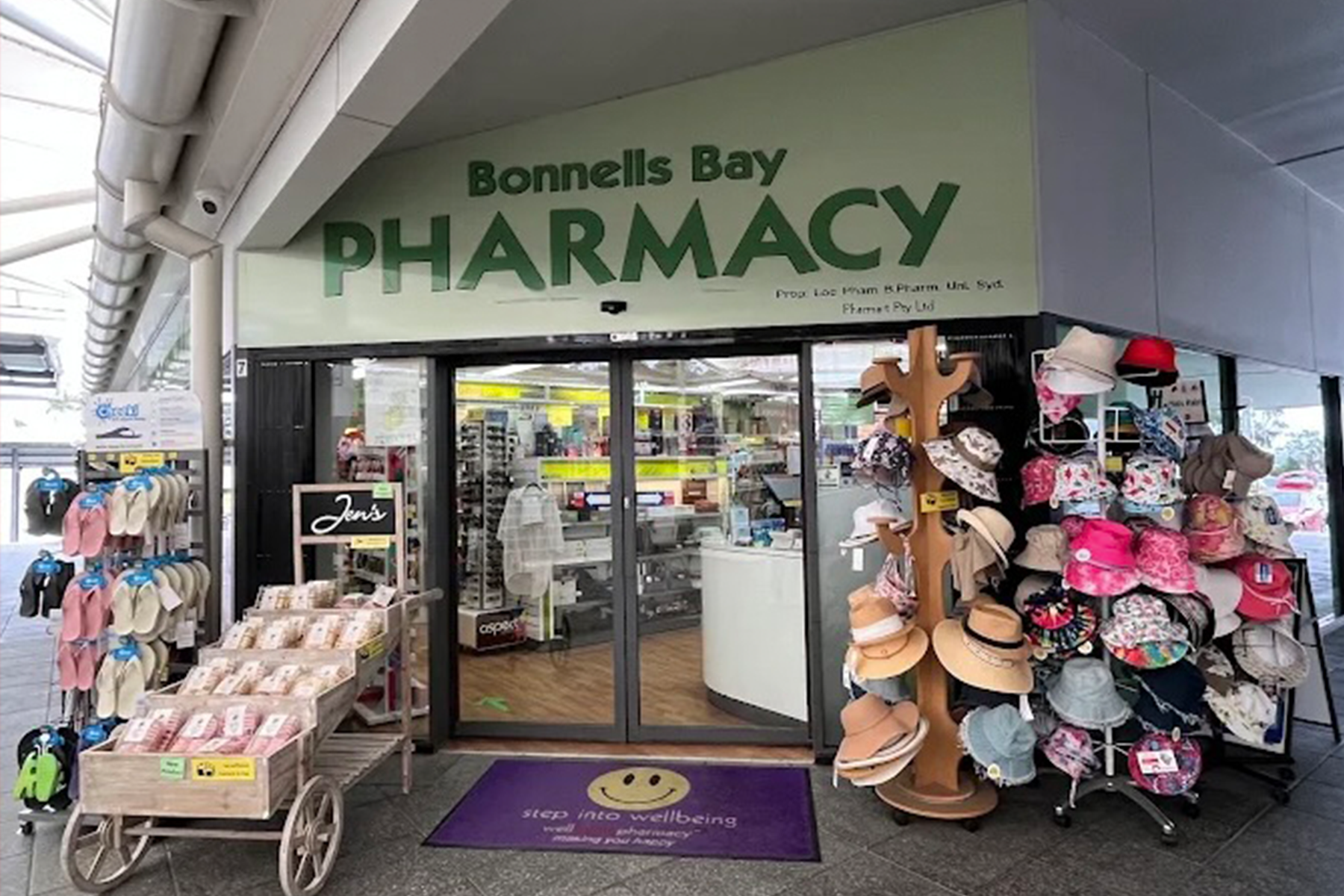 Entrance of Bonnells Bay Pharmacy with shelves of hats and slippers outside and store interior visible through sliding glass doors.