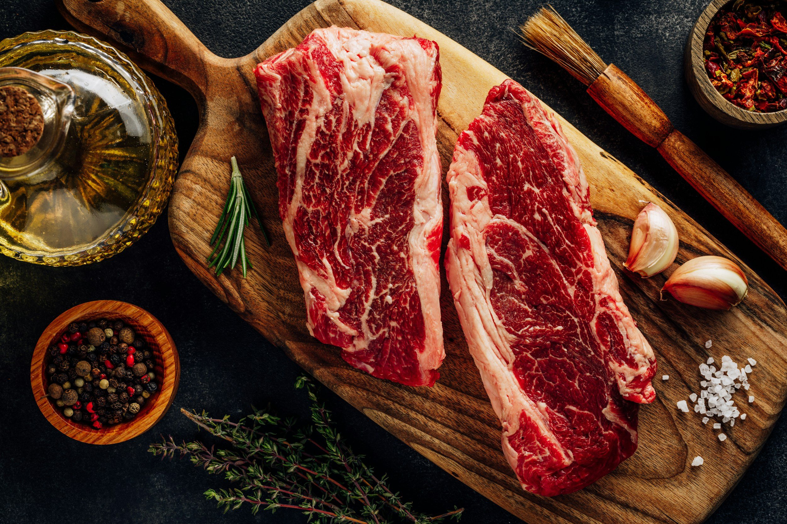 Raw steaks on a wooden cutting board with herbs, garlic, and spices around them.