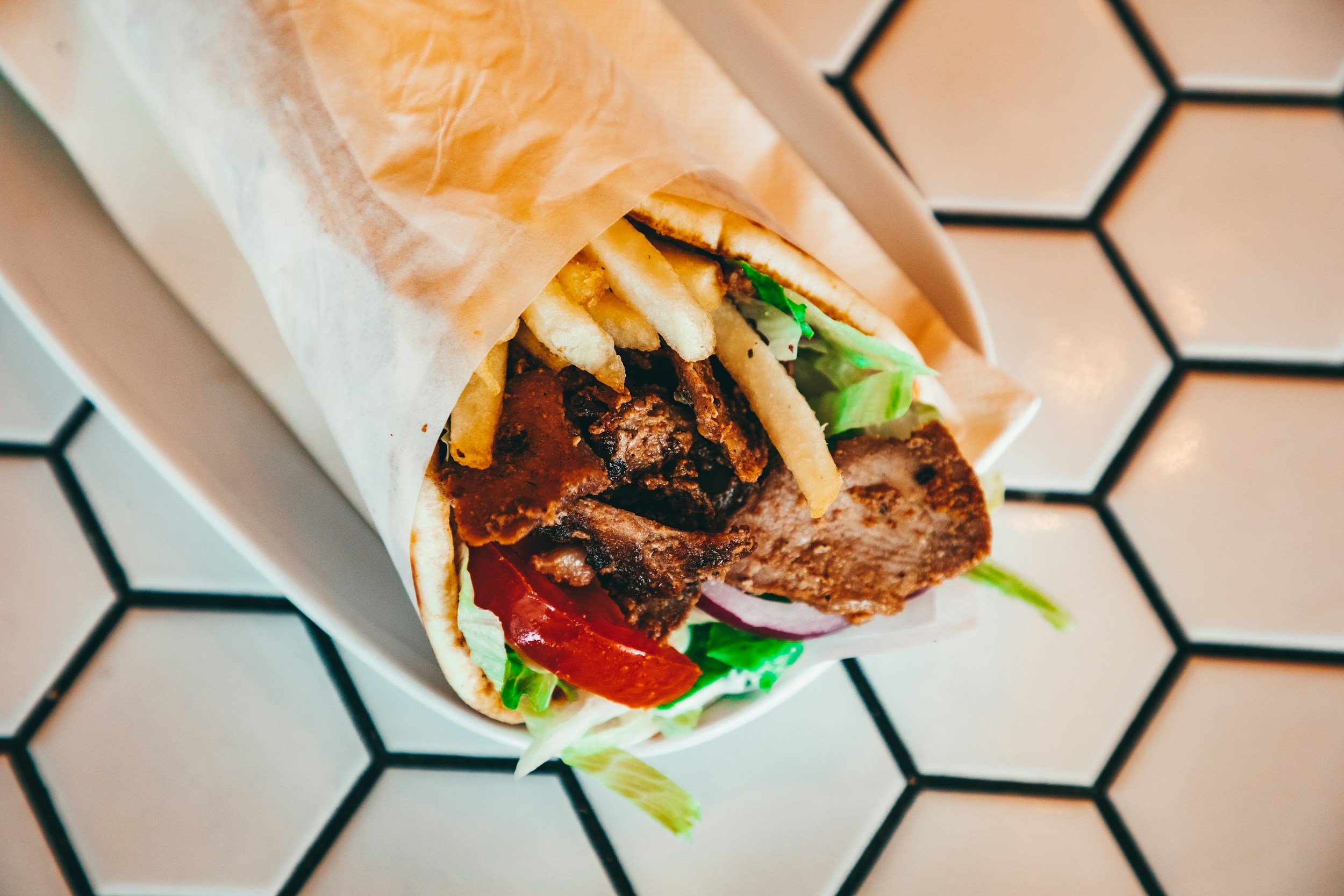 Close-up of a wrapped burger with lettuce, tomato, French fries, and beef patty on a white tray with a black honeycomb pattern.