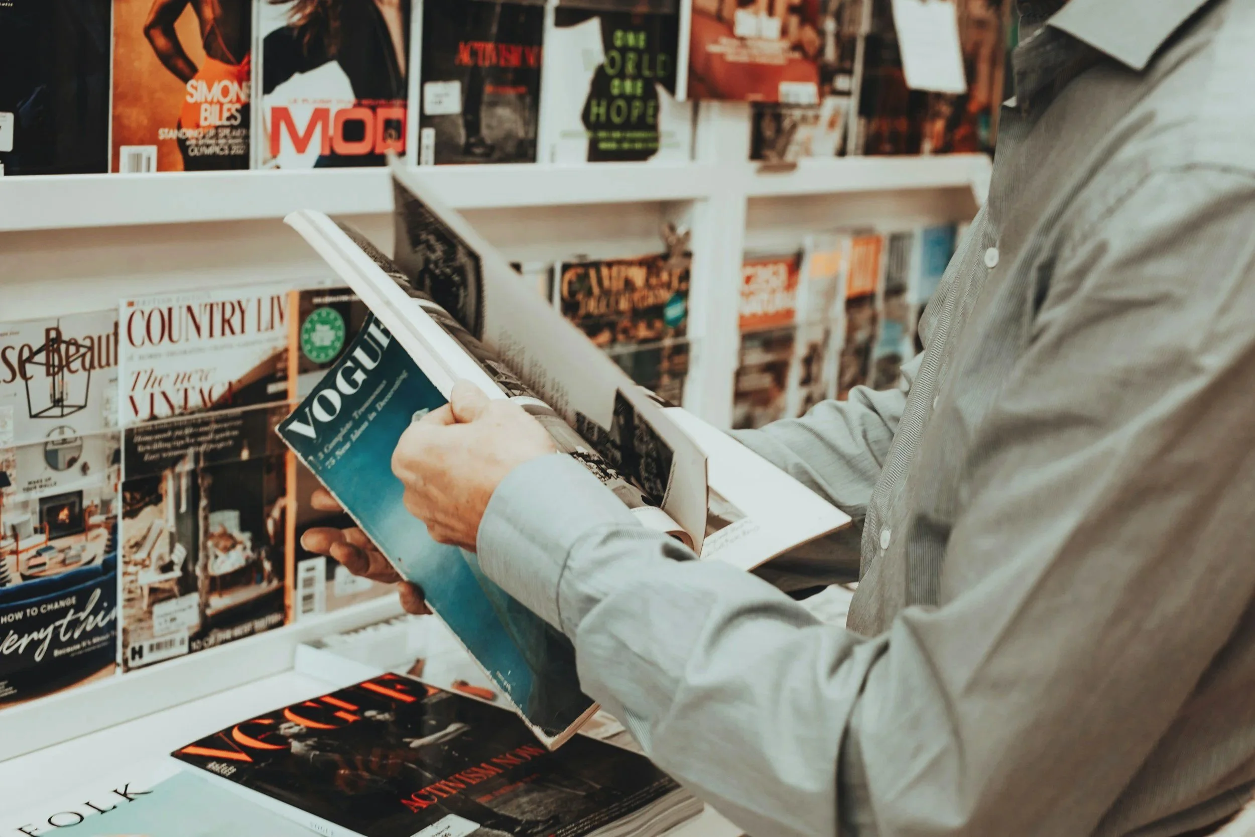 Person shopping for magazines in a store, holding a magazine titled 'Vogue'.