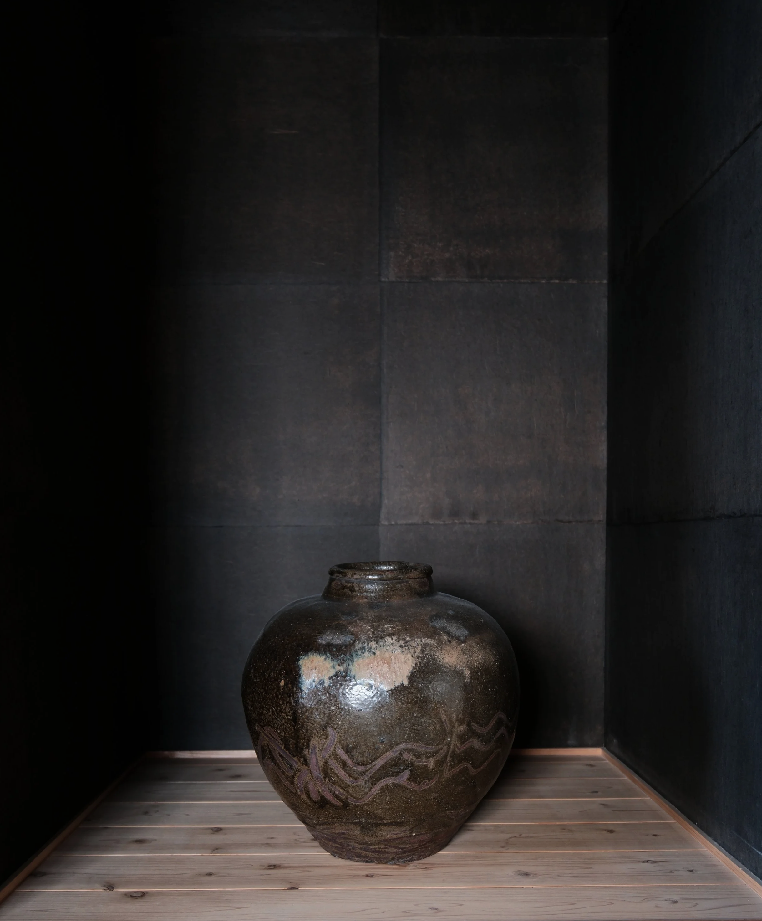 A large Japanese rustic brown ceramic vase with decorative patterns sits on a wooden floor against a dark, textured wall in a Japanese Ryokan.