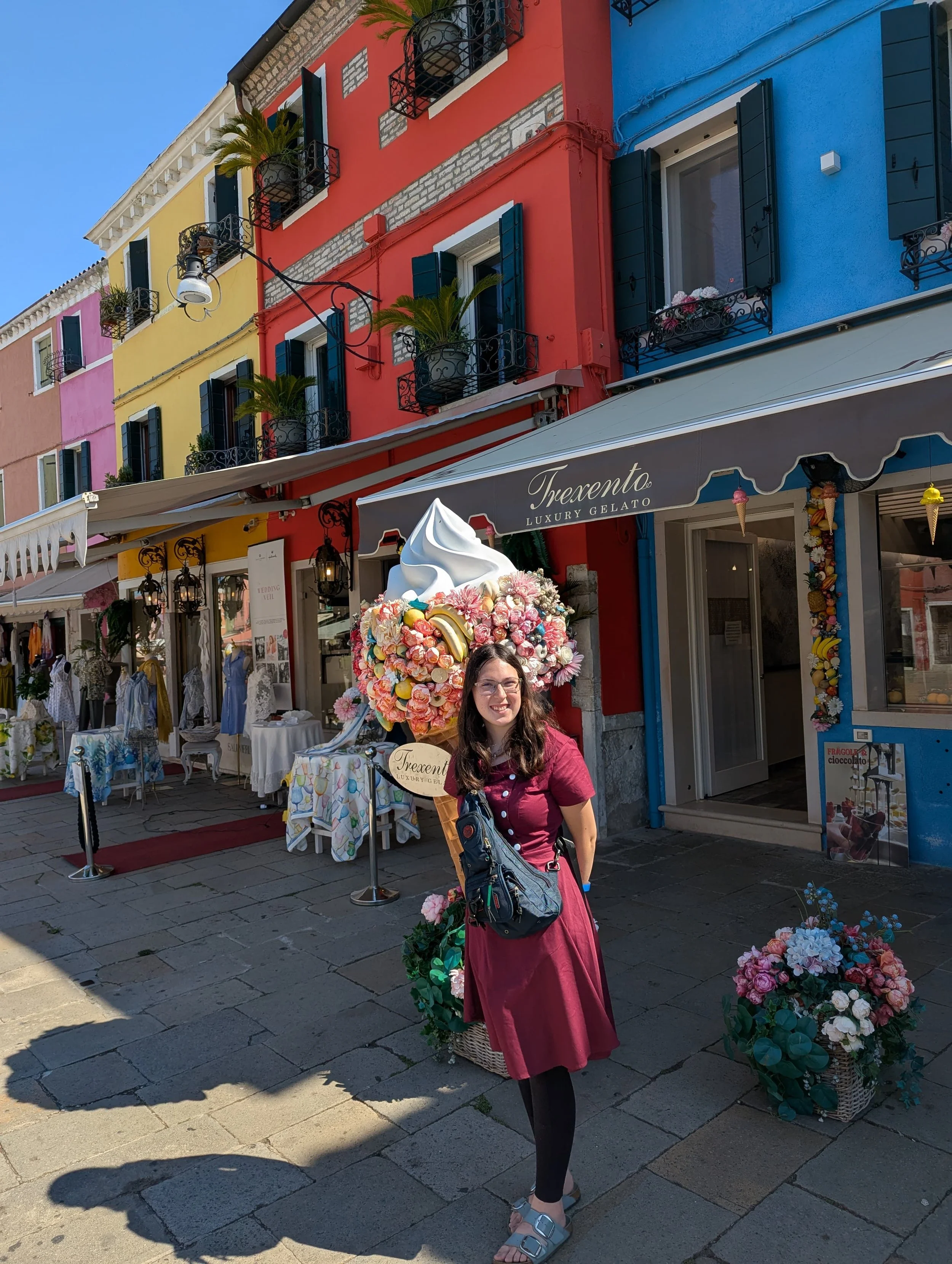 A woman with glasses and long dark hair stands on a street with colorful buildings in the background. She is smiling, wearing a long pink skirt, black tights, a maroon top, and sandals. She carries a small bag with a shoulder strap. There are flower arrangements and a sign for "Trecento Luxury Gelato" behind her. The street has a sidewalk with shaded and sunlit areas.