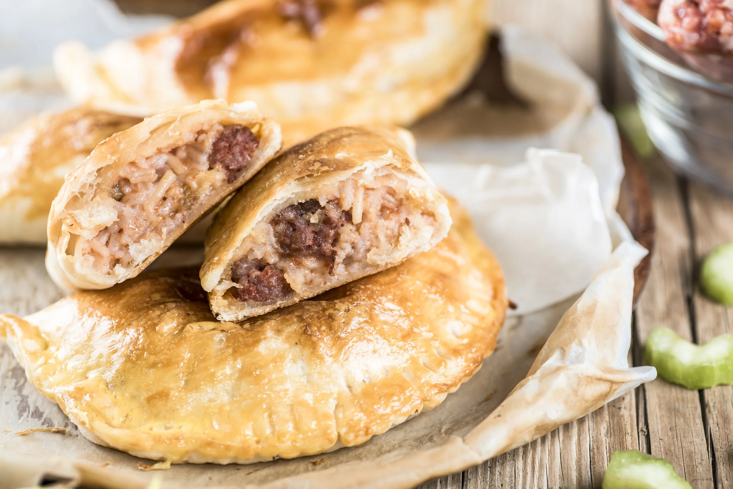 A close-up of a baked pastry stuffed with sausage, cheese, and herbs, with part of it cut open to reveal the filling. The pastry has a golden-brown crust and is placed on parchment paper on a rustic wooden surface, with a small container of meat or s