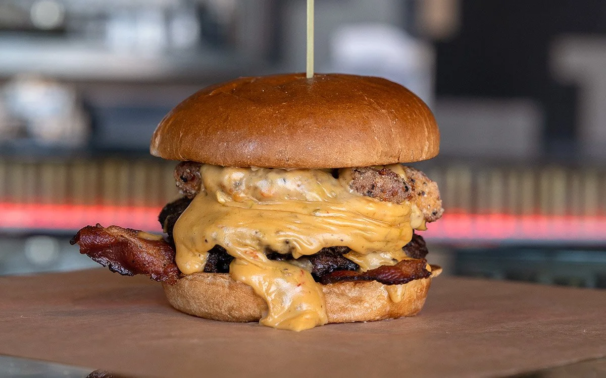 Close-up of a cheeseburger with bacon, melted cheese, and fried sausage patties in a brioche bun.