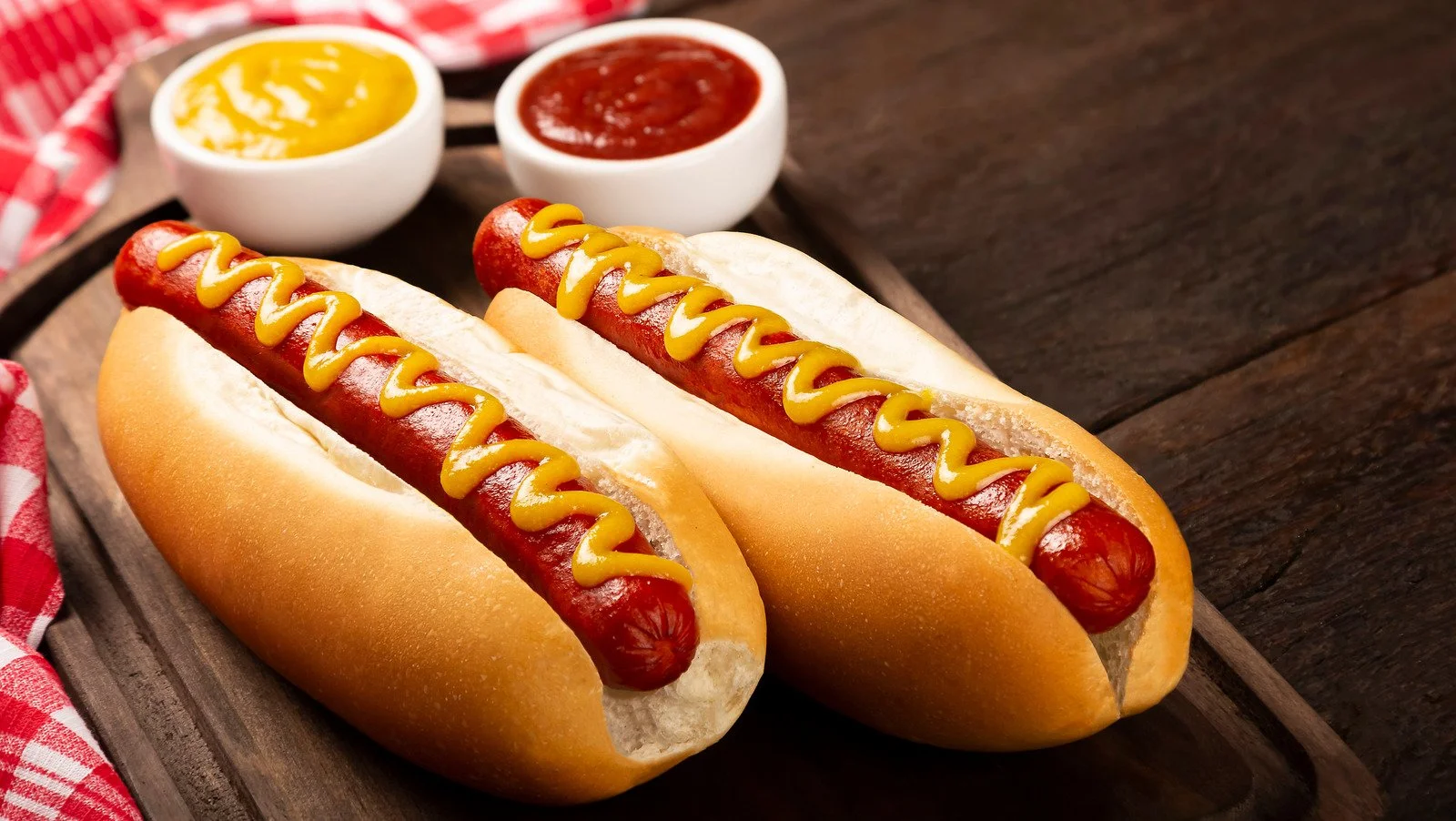 Two hot dogs in buns topped with mustard, placed on a wooden serving tray with two small bowls of mustard and ketchup in the background.