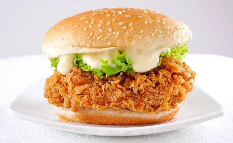 Fried chicken sandwich with lettuce, mayonnaise, and a sesame seed bun on a white plate.