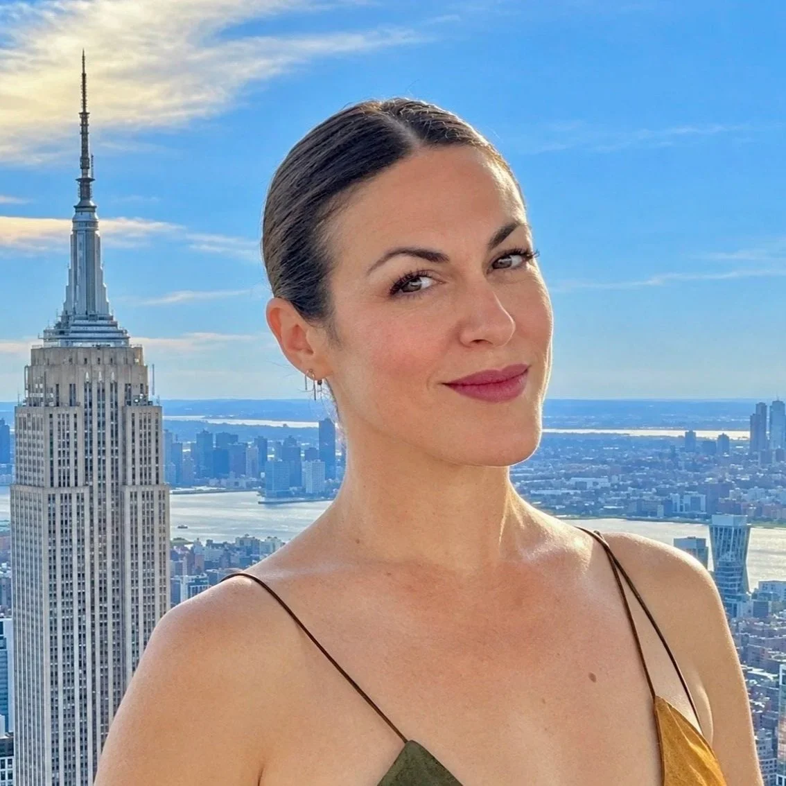 A woman with dark hair styled back, standing on a rooftop in New York City during daytime, with the Empire State Building in the background and a skyline view of the city and water.