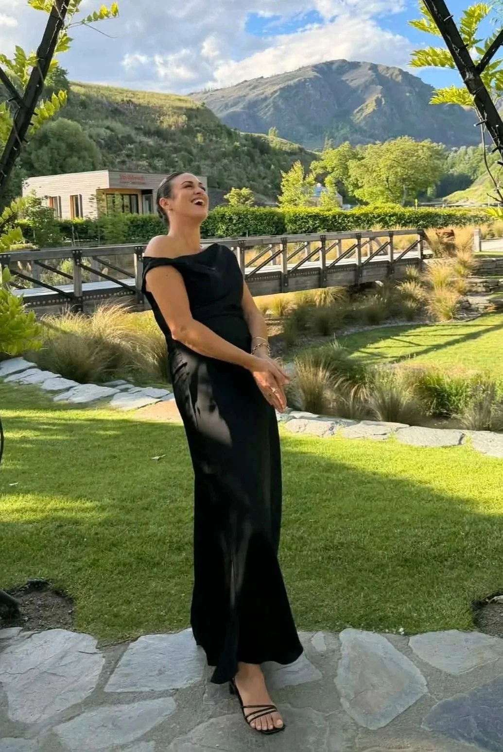 A woman in an elegant black dress standing outdoors on a stone pathway, smiling and laughing with mountains and trees in the background on a sunny day.