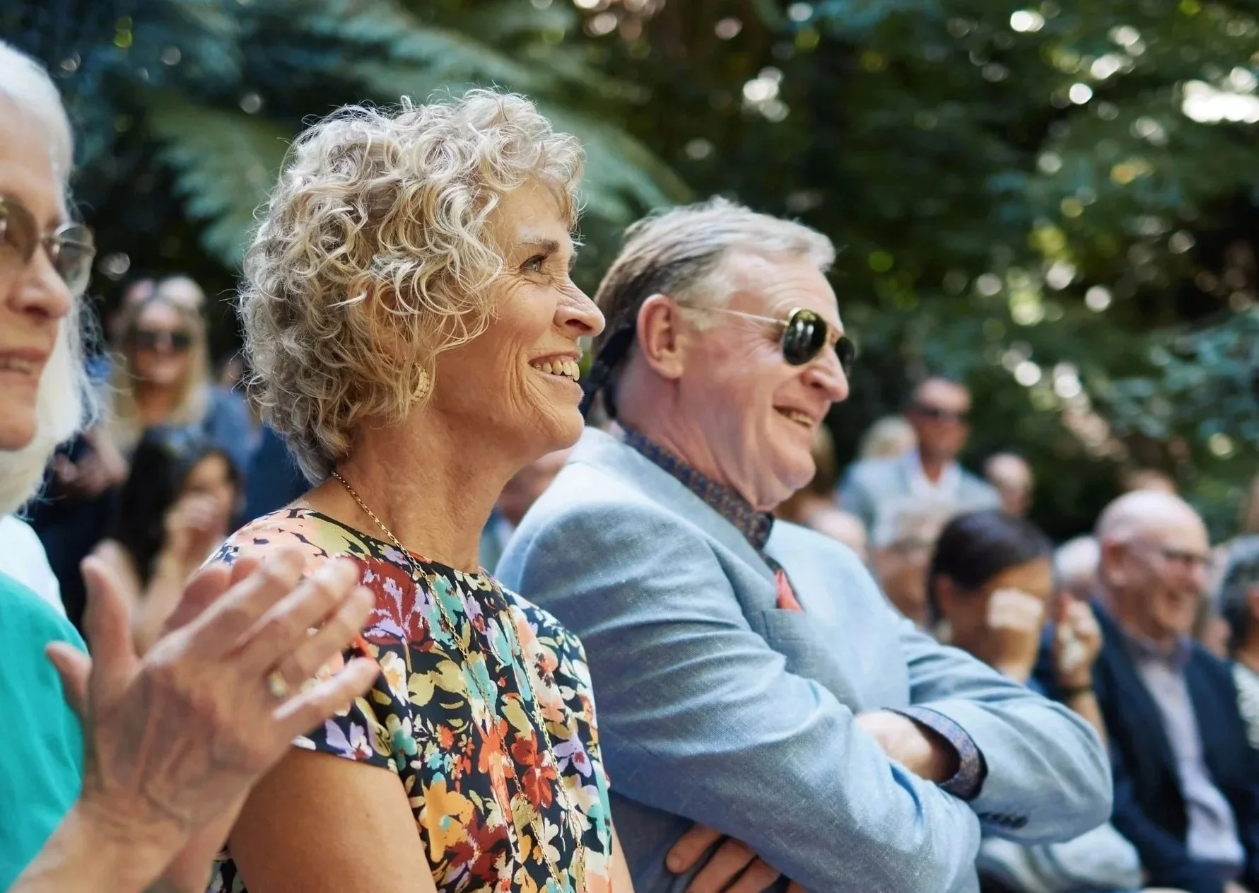 A group of people outdoors, sitting and smiling, with trees in the background. Focus on an elderly woman with curly blonde hair in the center, wearing a floral dress, and an elderly man with gray hair and sunglasses next to her.