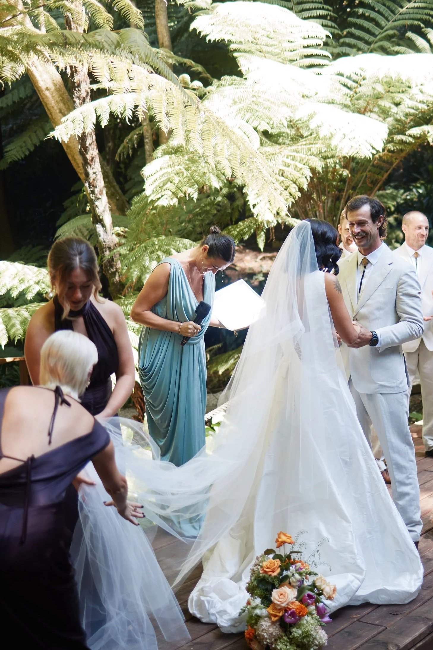 A wedding ceremony taking place outdoors in a lush, green, fern-filled setting. The bride, dressed in a white gown with a veil, is holding hands with the groom, who is wearing a white suit. Several guests are present, some of whom are assisting with or observing the ceremony. There are floral arrangements on the ground in front of them.