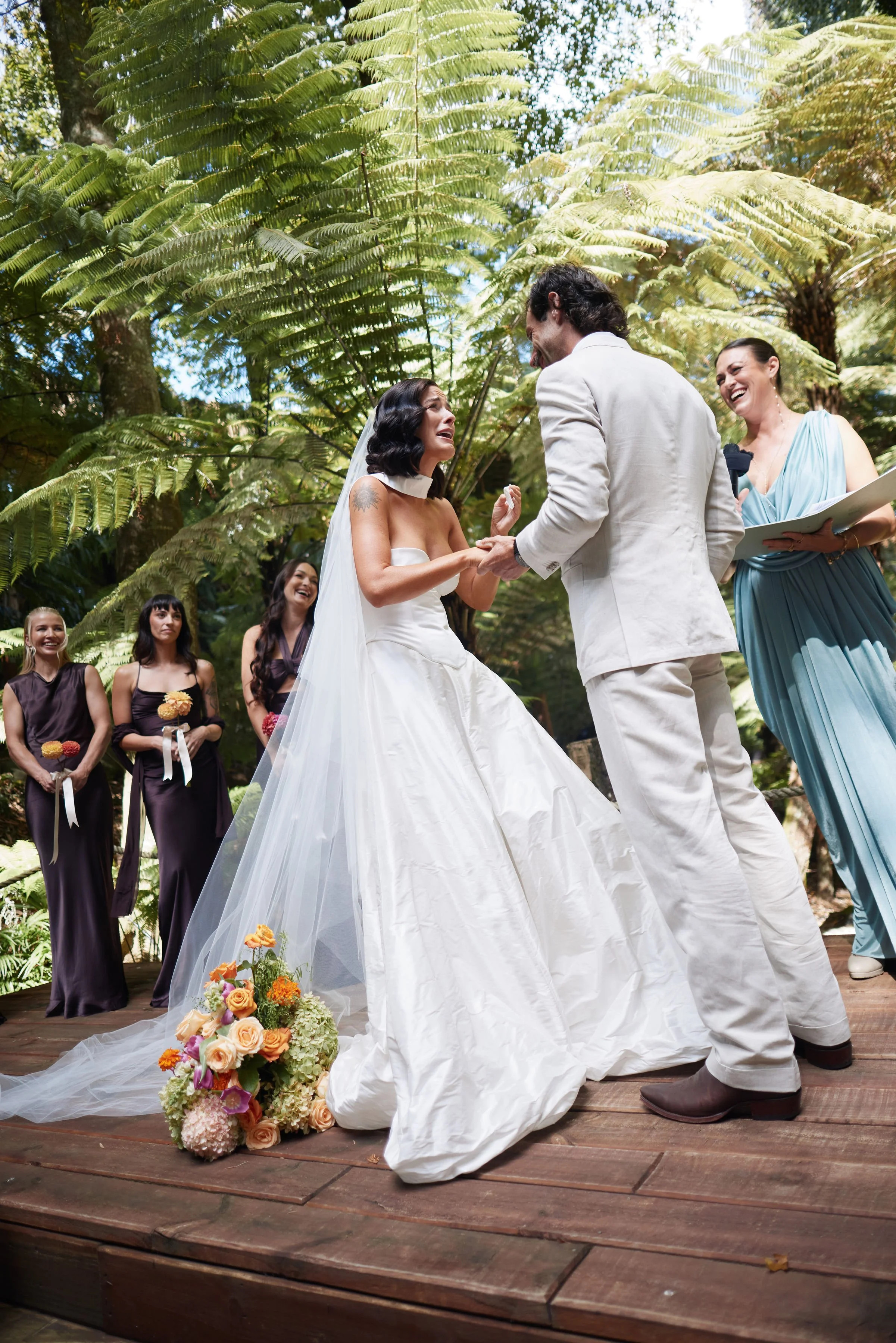 A wedding ceremony takes place outdoors beneath large green fern trees. A bride and groom hold hands and smile at each other, with the bride wearing a white strapless wedding dress and veil. A woman in a blue dress stands to the right, holding a microphone and a clipboard. In the background, four women in dark dresses watch and smile, some holding bouquets. A bouquet of flowers rests on the wooden floor in front of the couple.