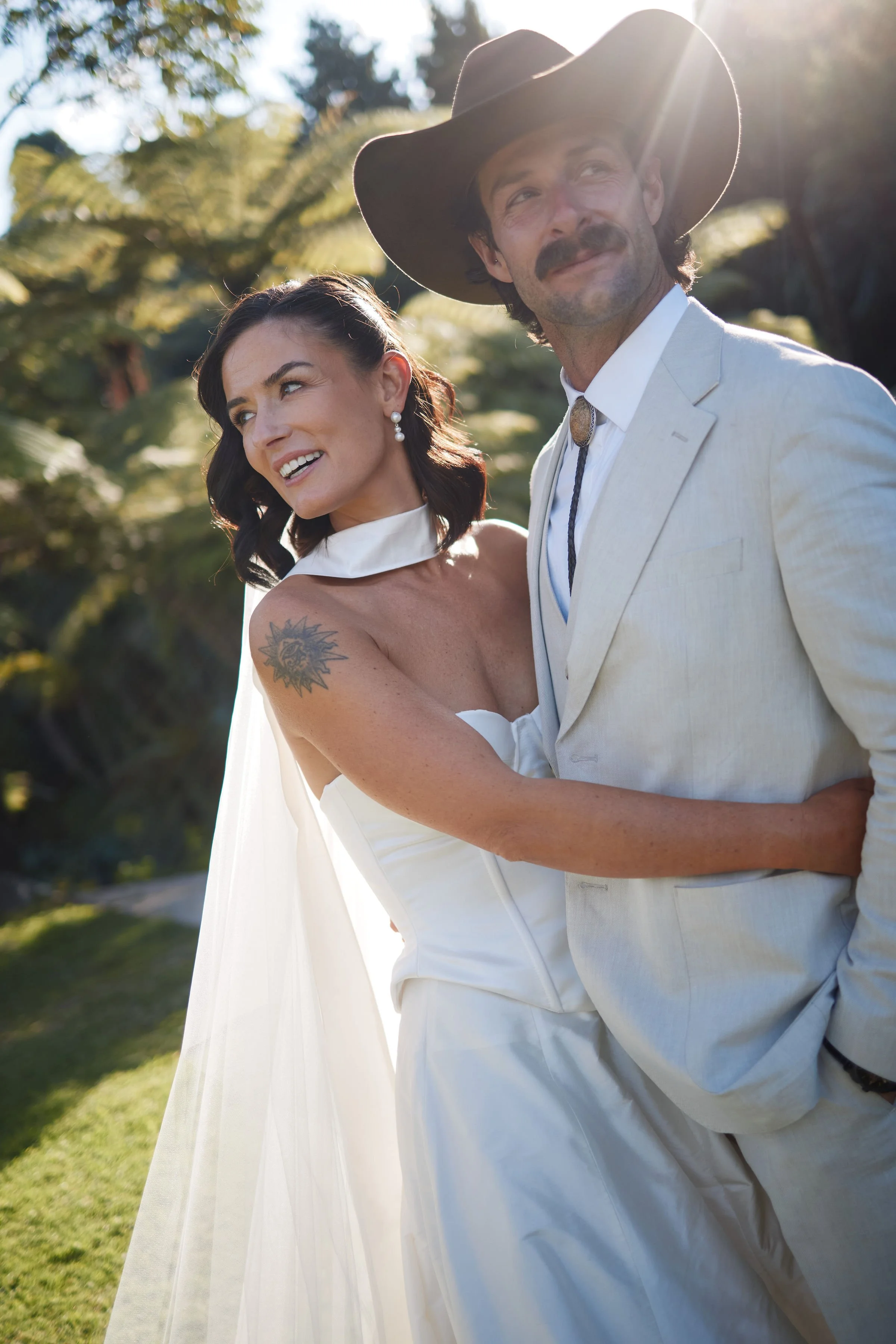 Aesha Scott and Scott Dobson on their wedding day, standing outdoors in a garden with green trees and sunlight in the background.