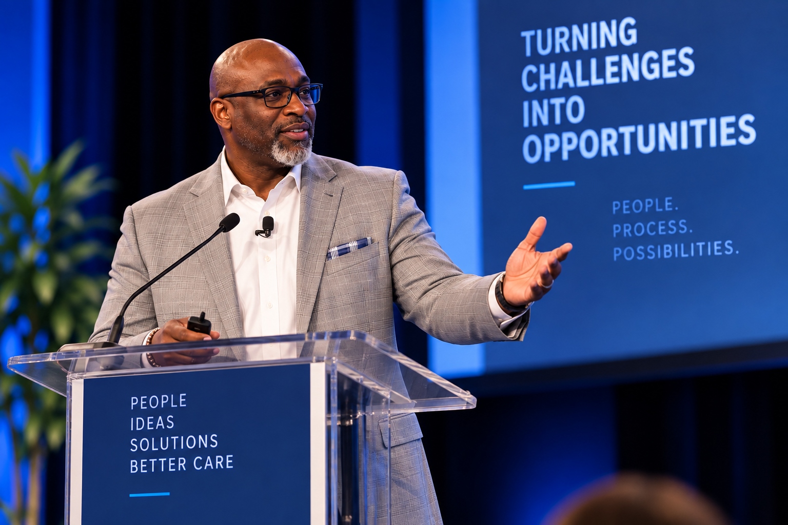 A man in a light gray suit and glasses giving a presentation at a conference, standing behind a clear podium with a blue sign that says 'People Ideas Solutions Better Care.' He is gesturing with his right hand, and there is a large screen behind him displaying the text 'Turning challenges into opportunities, people, process, possibilities.' There are microphones attached to his shirt and the podium, and a green plant is partially visible on the left side of the image.