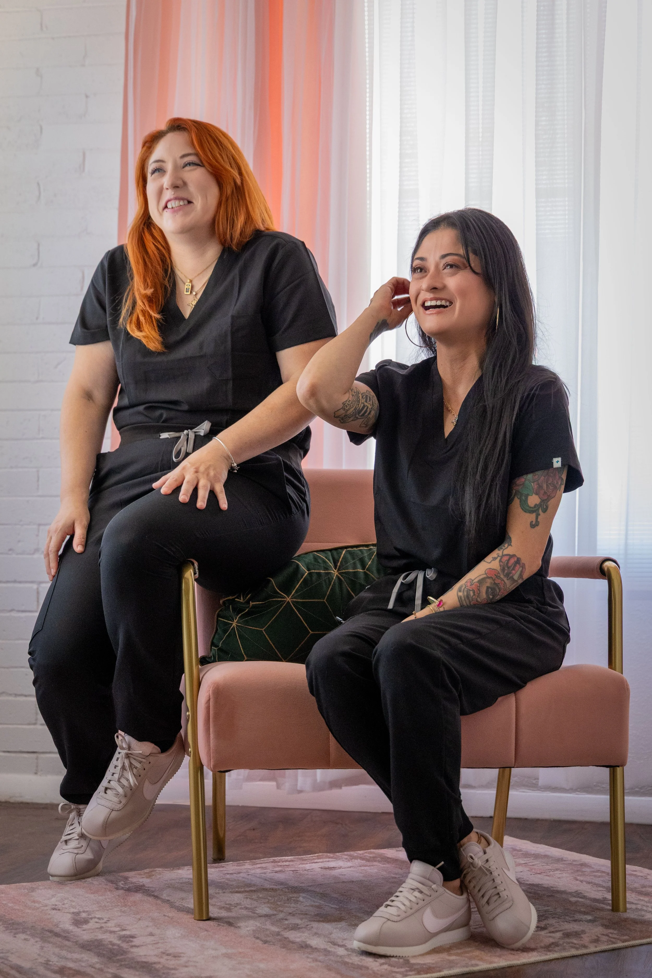 Two women in black medical scrubs sitting and standing on a pink chair in a room with white brick walls and floor-to-ceiling curtains, smiling and looking happy.