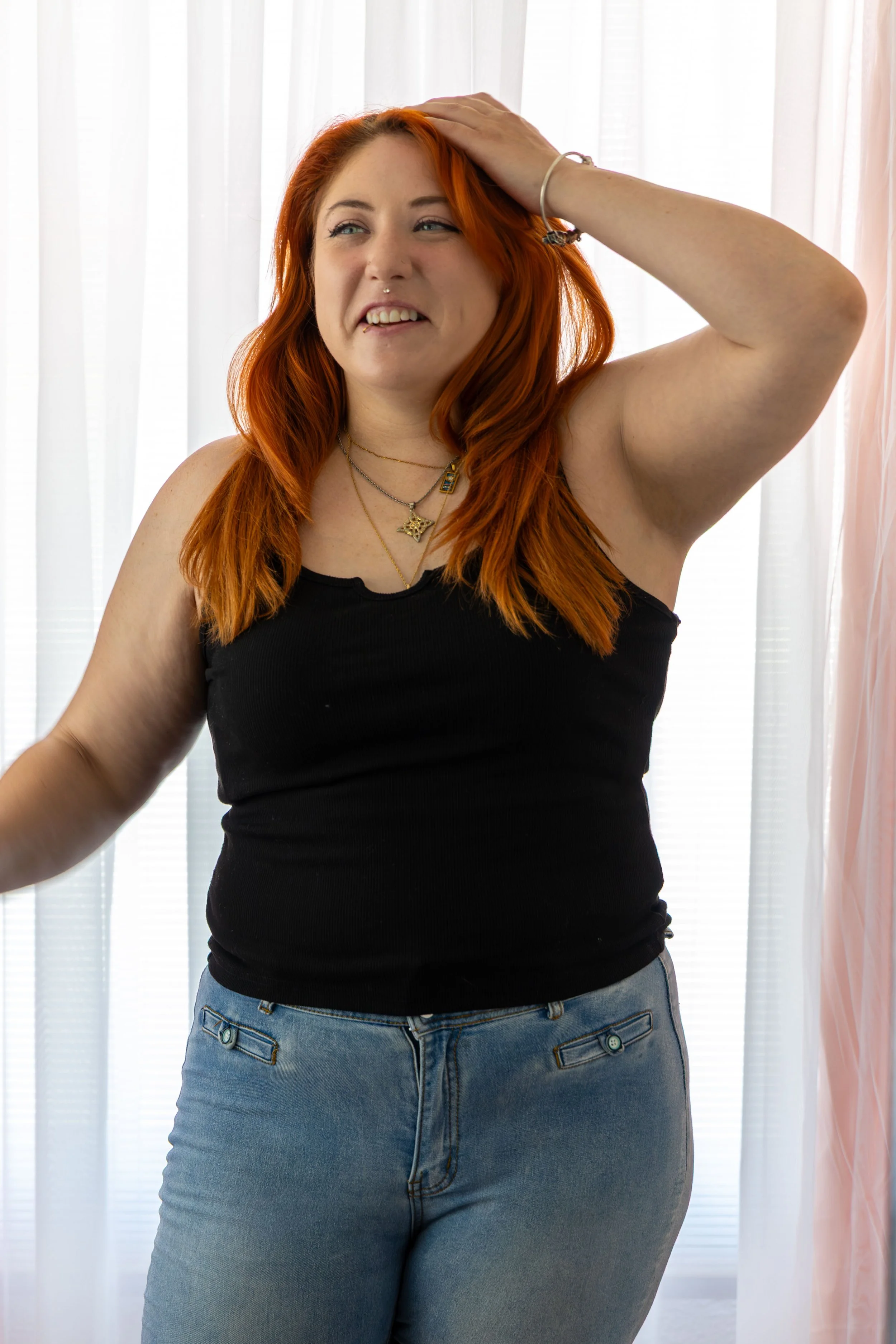 A woman wearing a black tank top and blue jeans standing in front of sheer white curtains, with her right arm raised and hand on her head, smiling slightly.