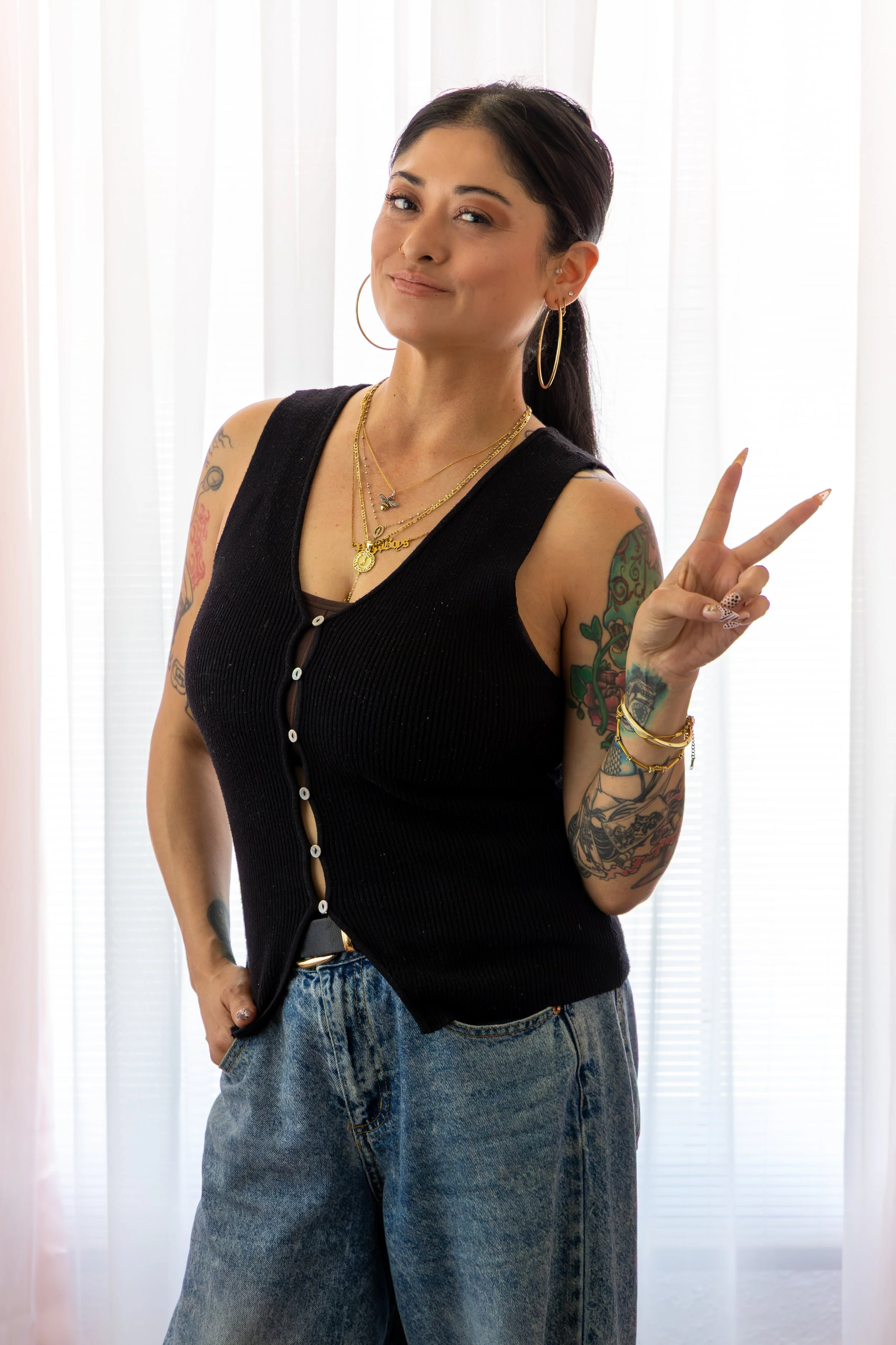 A young woman with dark hair and tattoos, wearing a black sleeveless top with buttons, blue jeans, large hoop earrings, and layered gold necklaces, standing in front of white curtains and giving a peace sign.