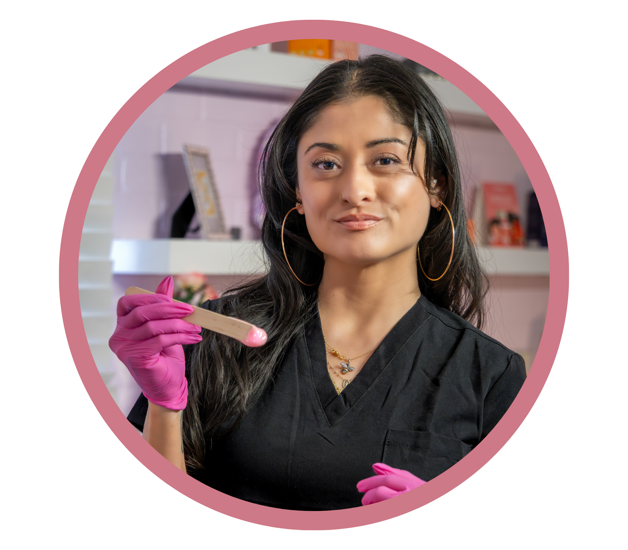 A woman with long dark hair and tattoos on her arm, wearing a black uniform and pink gloves, holds a swab inside a wax studio.