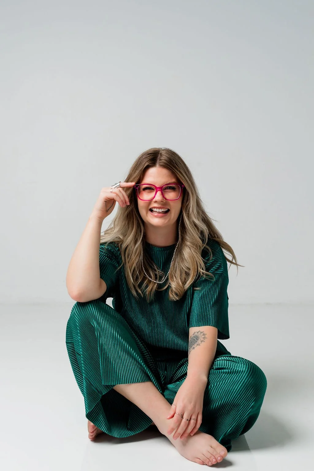 A young woman with long, wavy blonde hair, pink glasses, and a green striped outfit, sitting barefoot on the floor against a plain white background, smiling and adjusting her glasses.