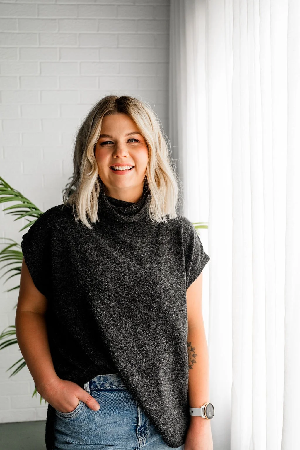 A smiling young woman with blonde hair wearing a dark gray turtleneck top and jeans, standing indoors in front of a white brick wall and sheer curtains, with a green plant partially visible behind her.