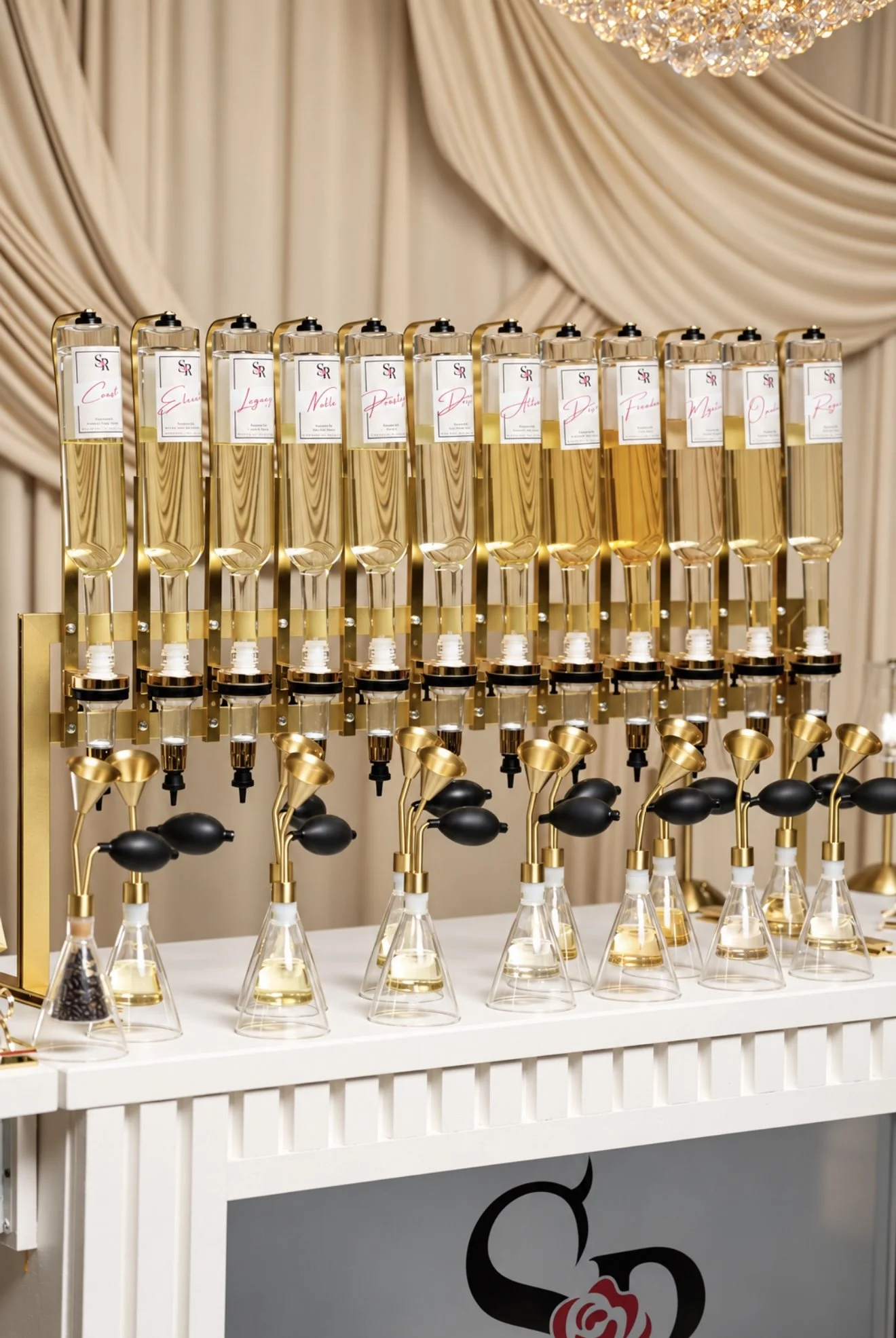 Elegant display of labeled perfume bottles and refill atomizers arranged on a white counter beneath draped fabric and a crystal chandelier