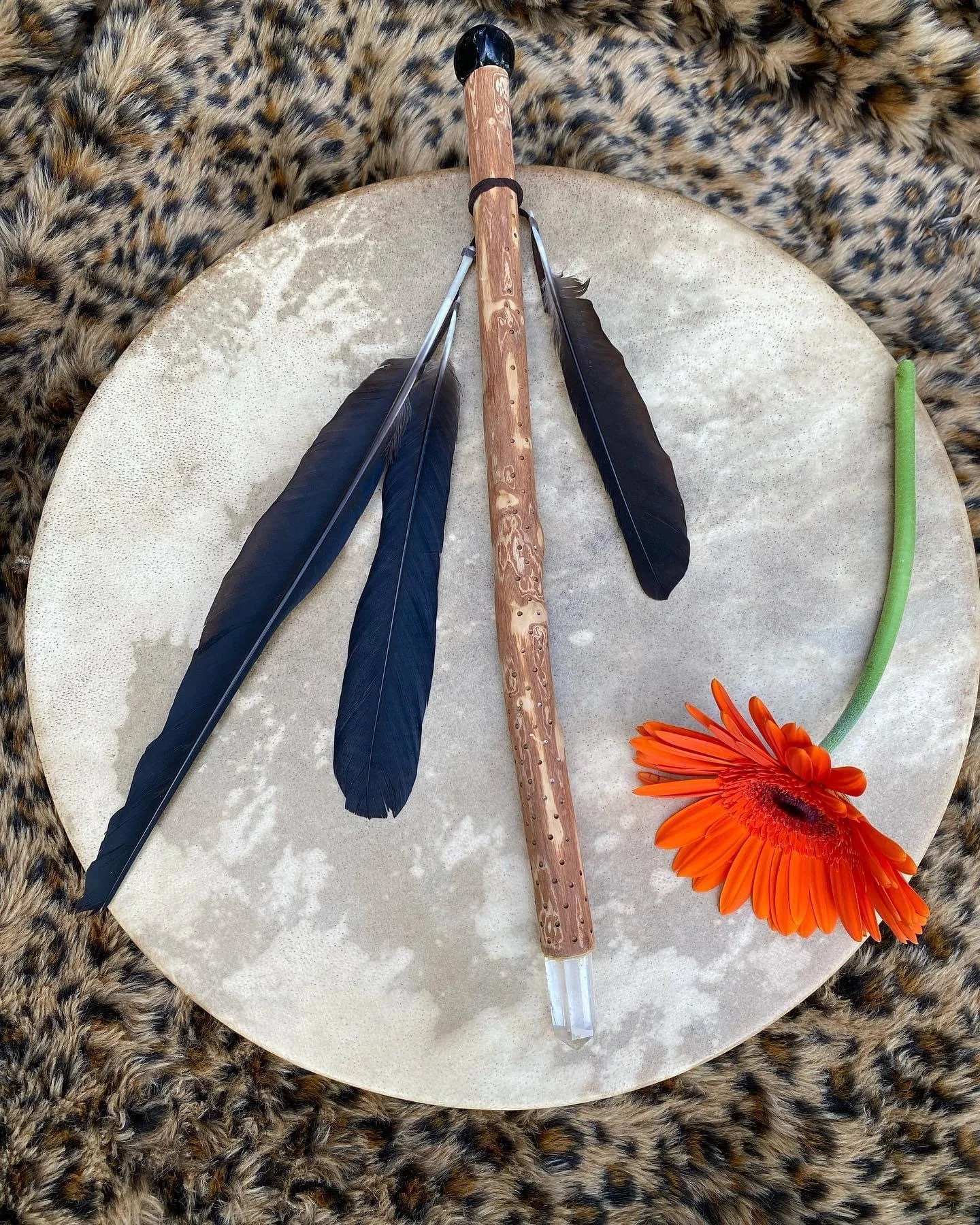 A white circular table with a wooden arrow, two black feathers, an orange flower, and green and orange decorations on it, with a leopard print fabric background.