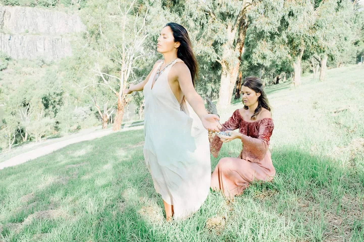 Two women in a grassy, wooded area outdoors, one standing with eyes closed and arm outstretched, the other kneeling with a plate, performing a ritual or blessing.