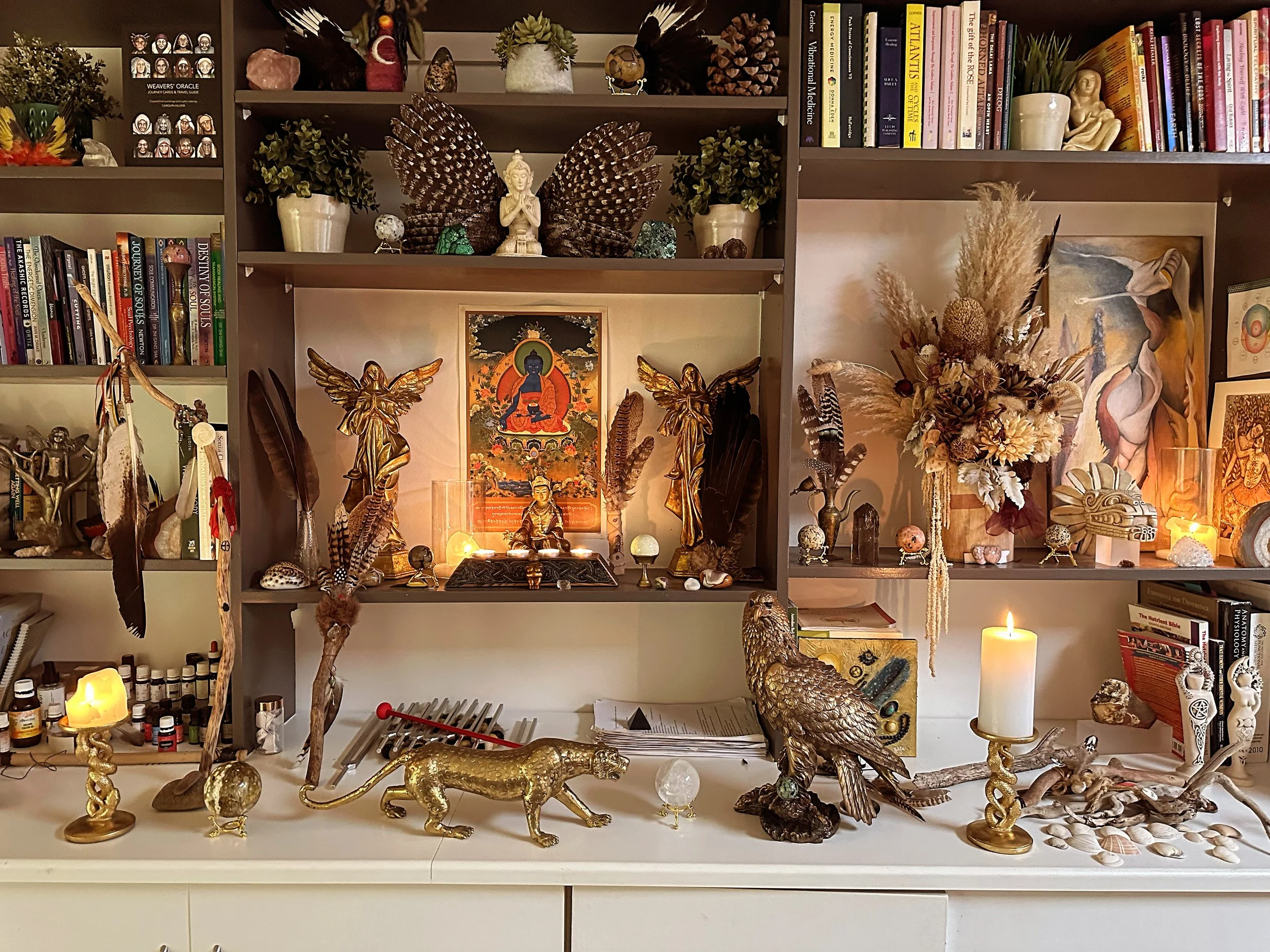 A decorative bookshelf filled with various spiritual and artistic items. The top shelf has plants, crystals, and other ornaments. The middle shelf contains books, some of which are related to healing and spirituality, along with sculptures and a framed picture. The bottom surface displays golden animal figurines, candles, shells, and natural elements like feathers and dried flowers, creating an eclectic and spiritual display.