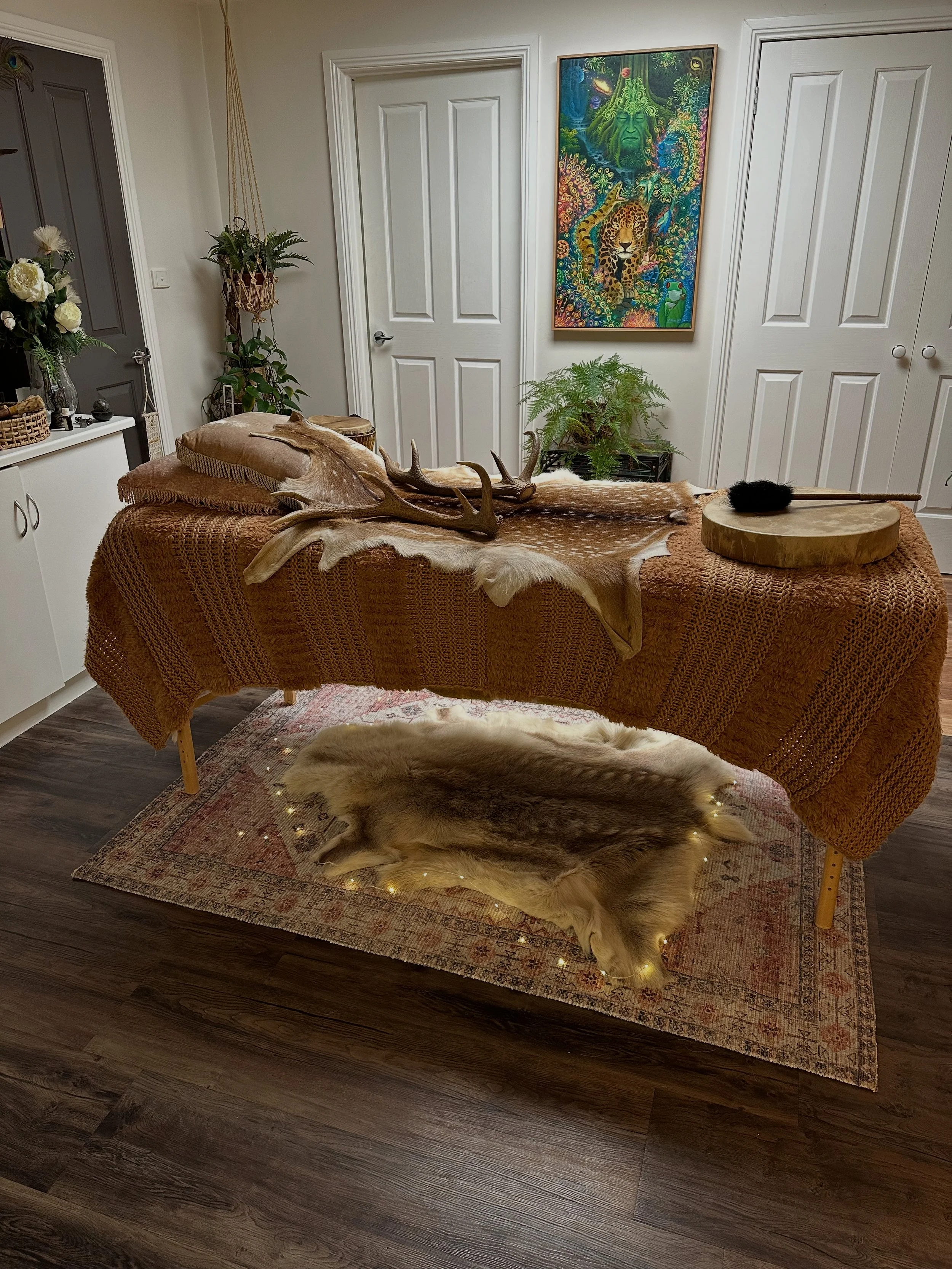 An indoor room with a massage or treatment table covered with a brown-orange woven blanket, a deer hide with antlers, and a fur rug underneath. There are pillows on the table, a stick with black fluffy object resting on a round wooden platform, string lights on the floor, potted plants, and a colorful jungle-themed painting on the wall.