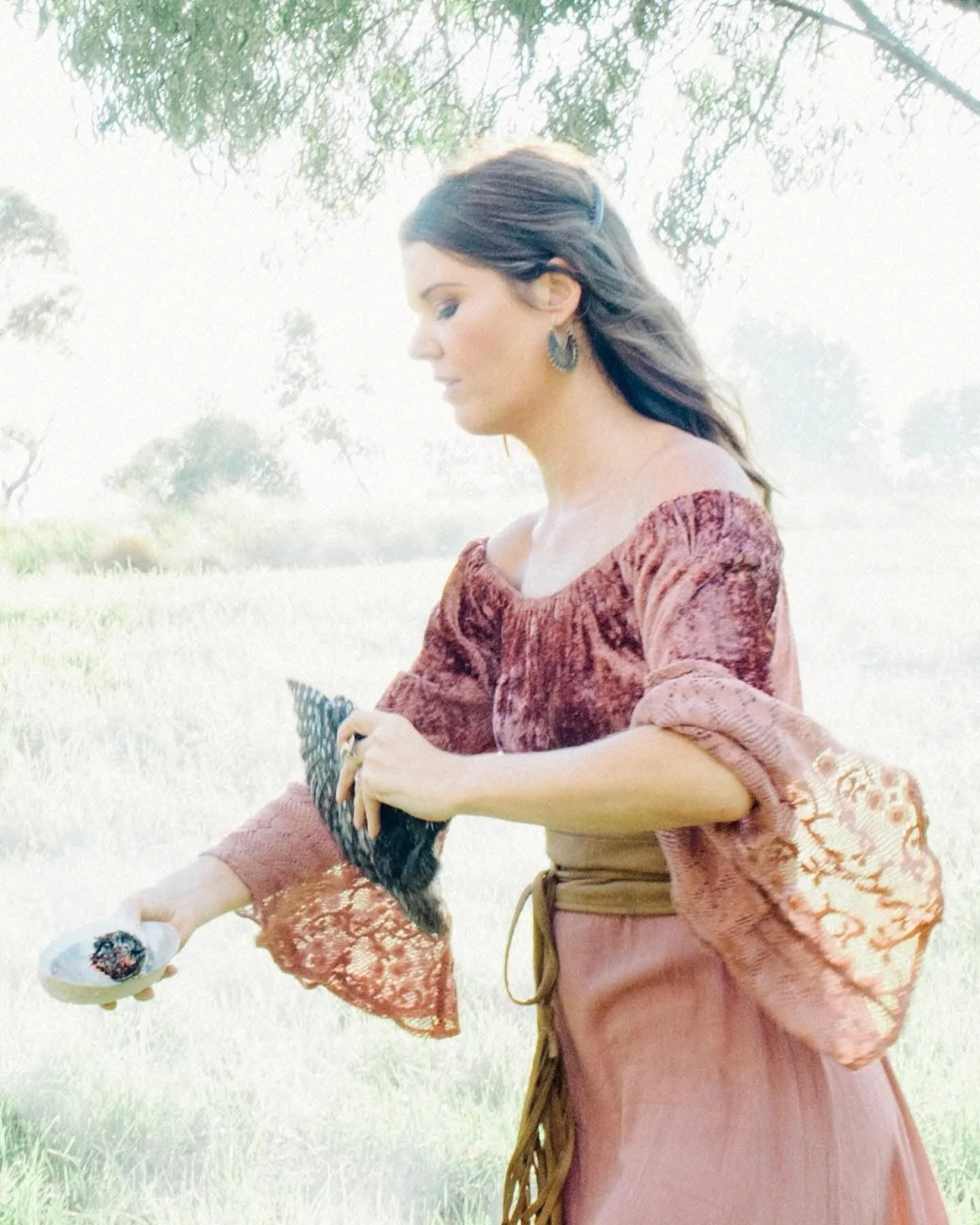 A woman in vintage-style clothing sitting outdoors in a grassy field, holding a small dish with a cupcake, with trees in the background.