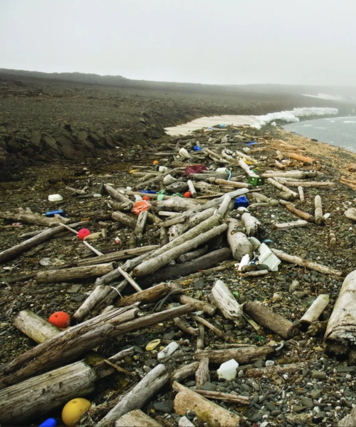 A beach with dark rocks and driftwood, littered with trash including plastic bottles, foam containers, and floats, near the ocean with waves and a cloudy sky.