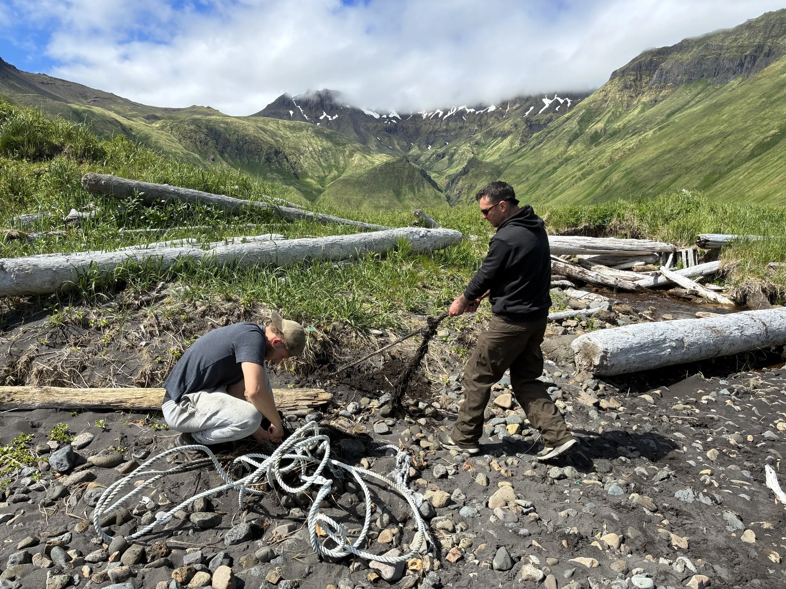 Two men working outdoors near a rocky stream, with green hills and mountains in the background under cloudy skies.