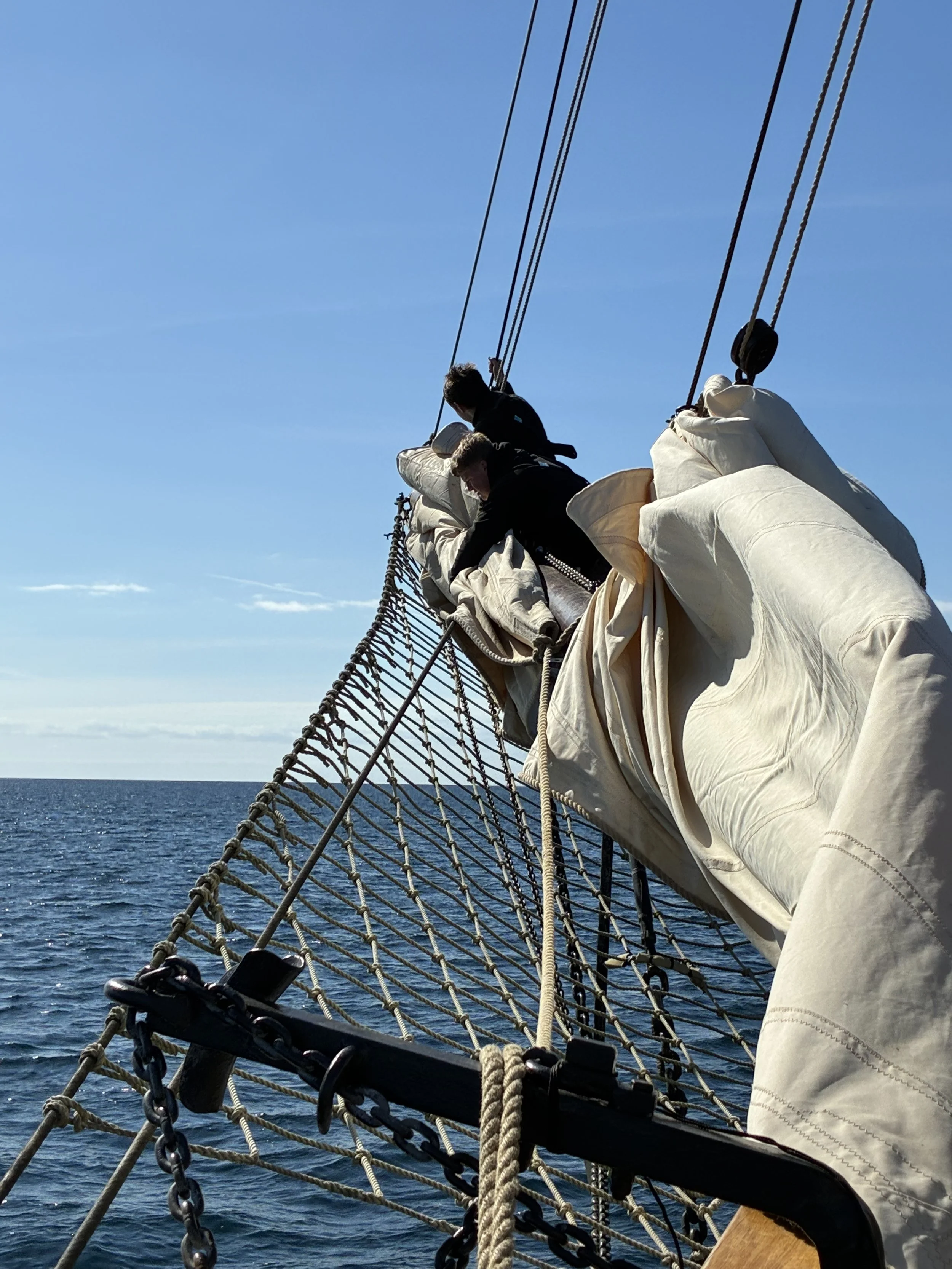 View of a ship's rigging and sails from the deck, with the ocean and sky in the background.