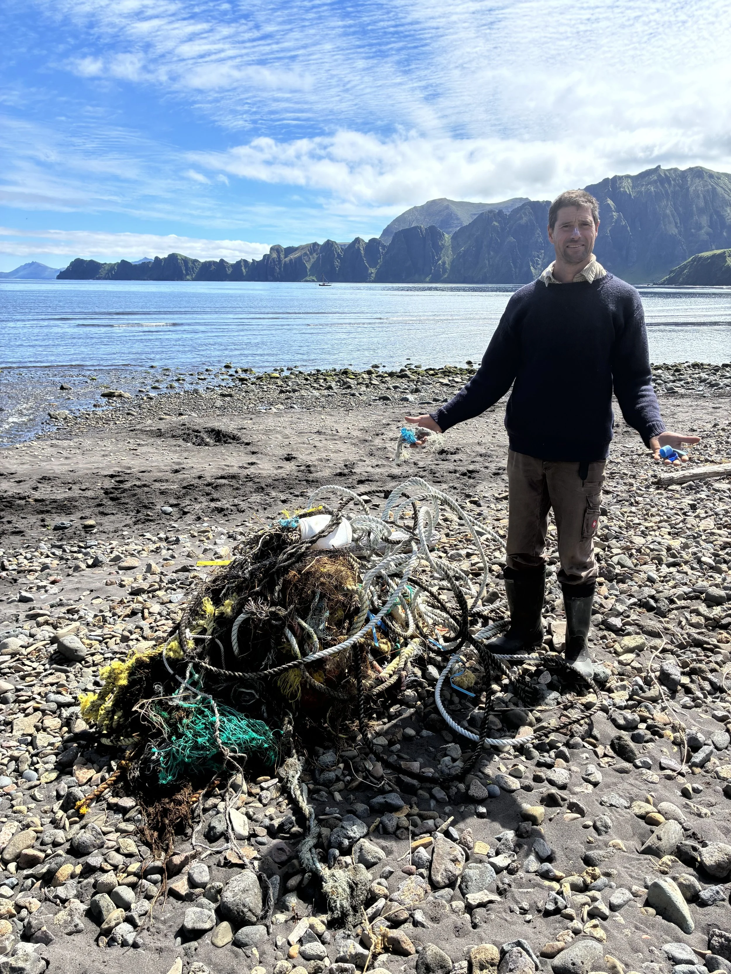 A man standing on a rocky beach with mountains and a blue sky with clouds in the background, holding ropes and surrounded by tangled fishing nets and debris.