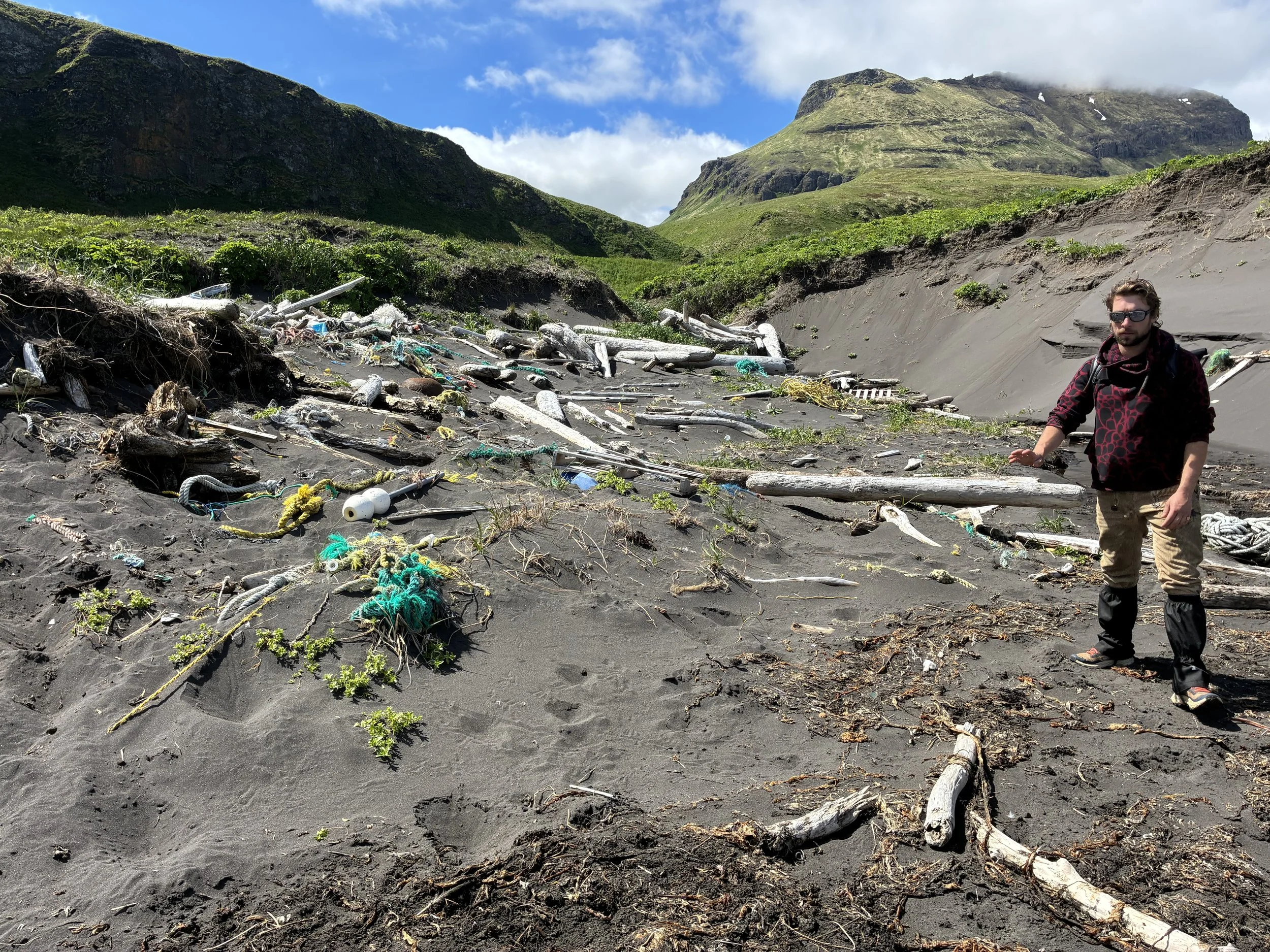 A man wearing sunglasses and outdoor gear stands on a black sand beach strewn with driftwood and debris, with green hills and mountains in the background under a partly cloudy sky.