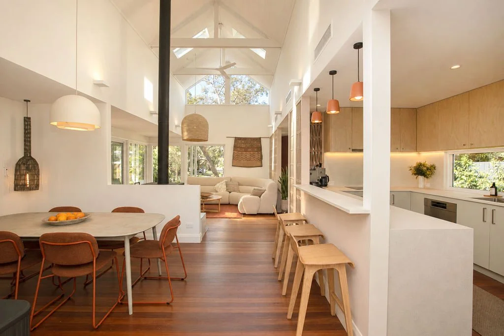 Yaraana Pearl beach holiday Luxury house Open-concept living room and kitchen with high vaulted ceiling, wooden floors, white walls, and large windows showing outdoor trees.