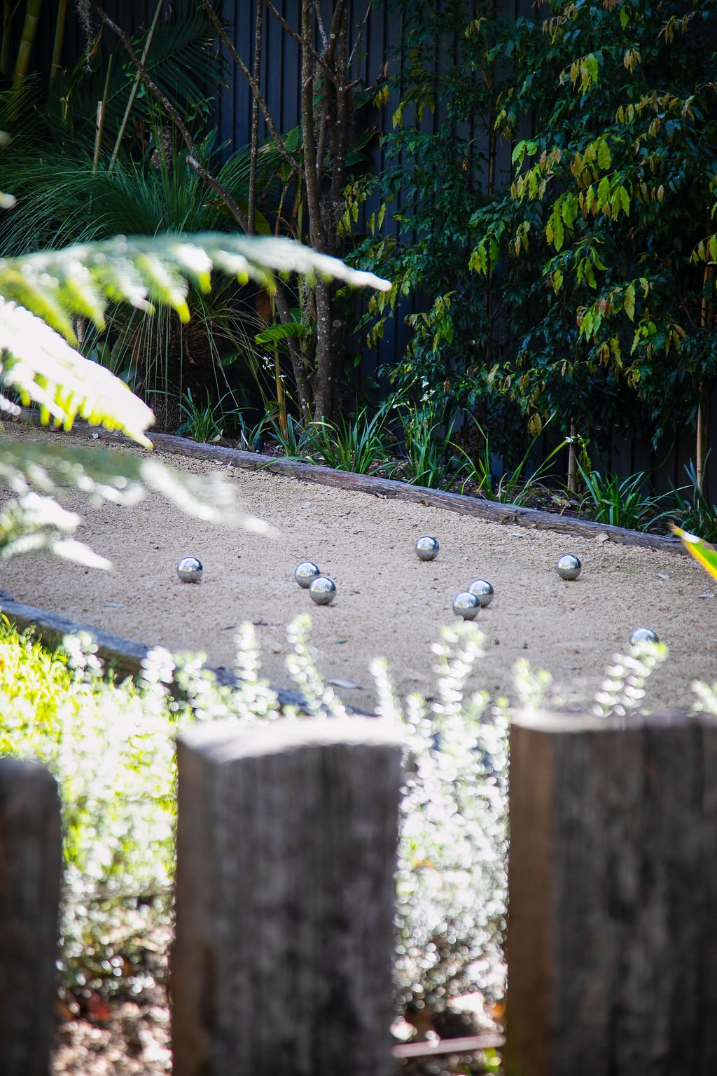 A sand-colored bocce court with metal balls scattered on it, surrounded by lush green plants and trees.