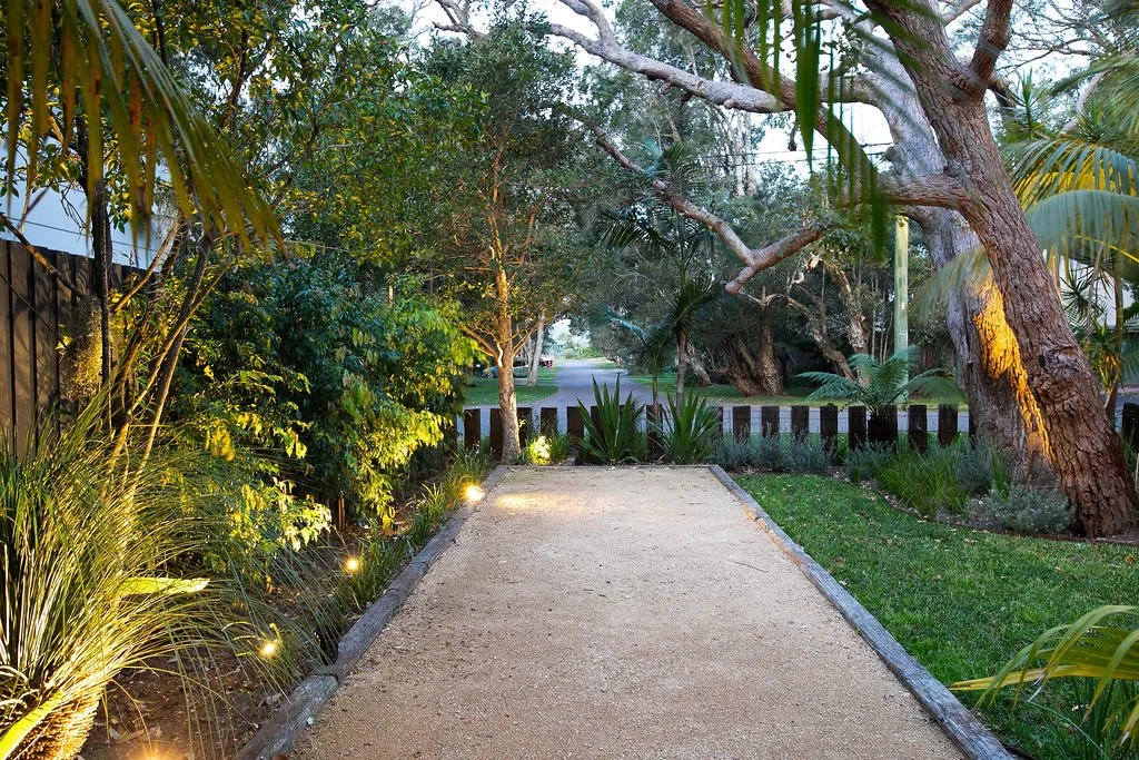 A gravel pathway lined with small lights on the left side, surrounded by lush green tropical plants and trees, leading to a street with parked cars and greenery in the background during twilight.