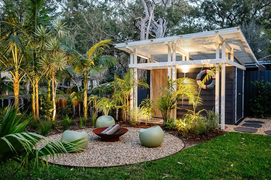 A modern backyard garden with tropical plants, outdoor seating, fire pit, and a small dark-colored building with a white roof and a life preserver decoration.