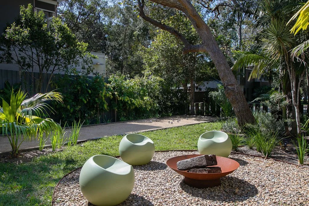 Sunny backyard with three modern white chairs, a fire pit filled with logs, surrounded by grass, rocks, trees, and lush plants.