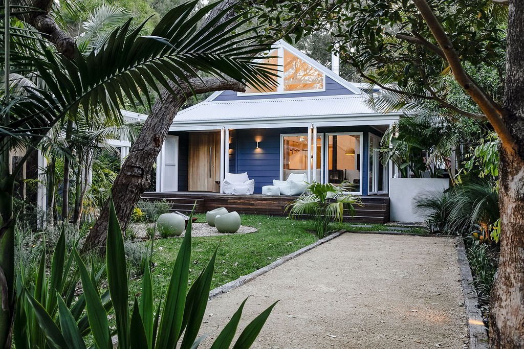 A modern blue house with large glass doors opening to a patio, surrounded by lush greenery and trees, with a gravel pathway leading to the house.