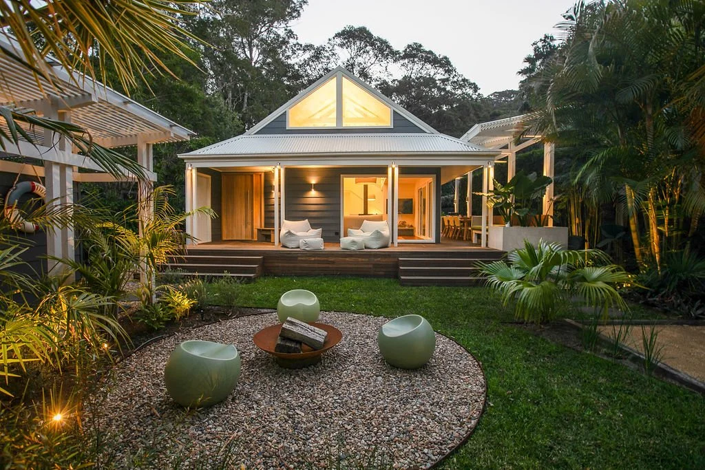 A modern house with a porch and outdoor seating in a lush garden at dusk, illuminated by warm interior light.