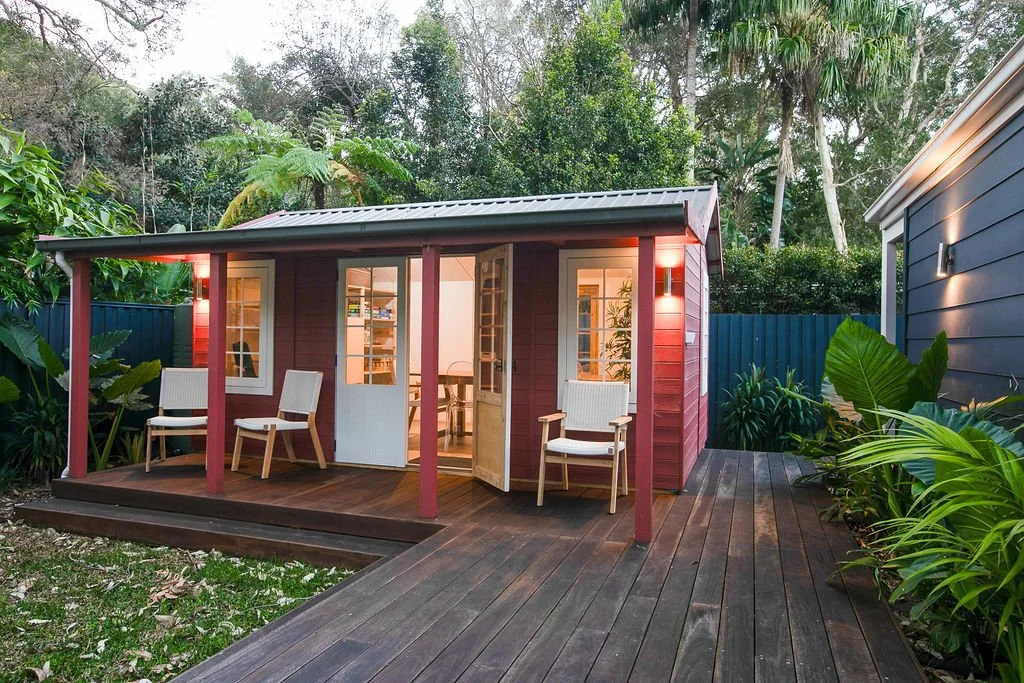 A small red wooden house with white-framed windows and a door located at the center, surrounded by lush green tropical plants, with a front porch furnished with two white chairs and a wooden deck, in a backyard setting with tall trees in the backgrou
