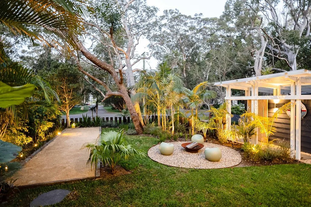 An outdoor garden at dusk with a sandy pathway, lush green plants, and a large twisting tree. There are decorative white spheres and a wooden tray on a gravel circular area. A modern white pergola with lighting is on the right.