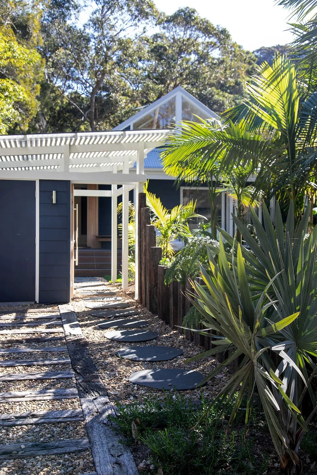 A modern house with a white pergola, surrounded by lush green plants and trees, with a gravel pathway leading to the entrance.