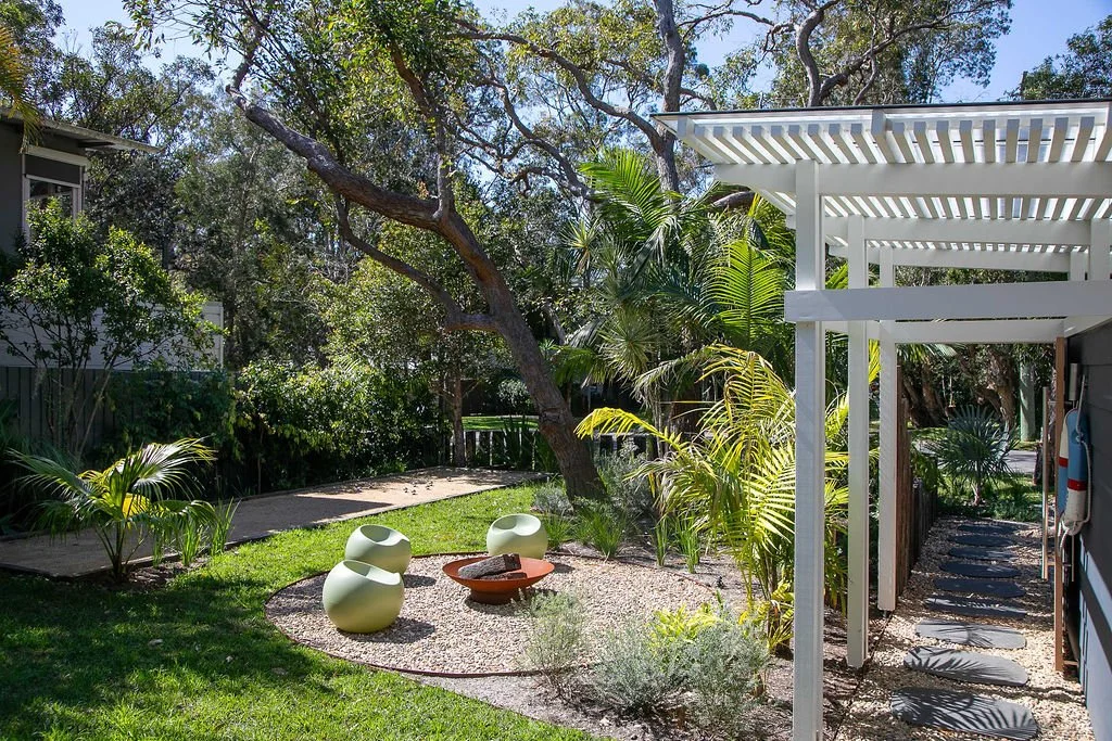 A backyard garden with large trees, tropical plants, and modern white garden chairs surrounding a fire pit, with a white pergola shade structure on the right.