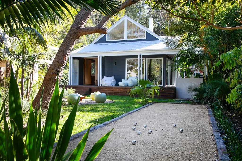 A backyard with a kerb ball court, lush greenery, and a modern house with large glass doors and a small deck.