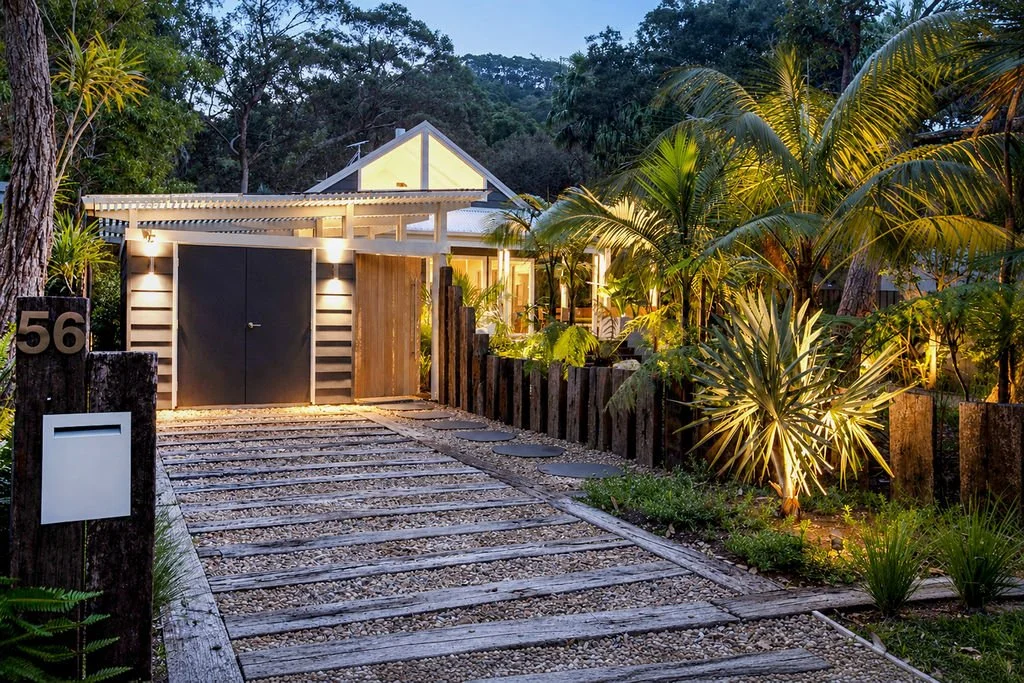 House with a sandy pathway, surrounded by tropical plants and trees, with outdoor lighting illuminating the garden area during dusk or nighttime.