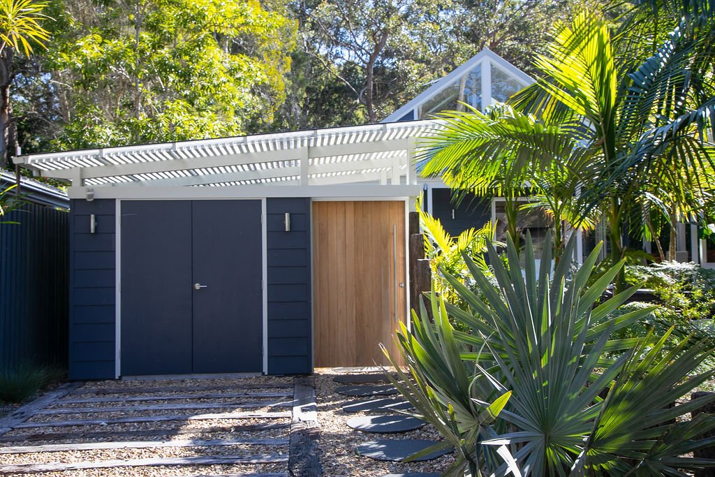Modern backyard with wooden and metal fencing, gravel pathway, lush tropical plants, and a glass-roofed greenhouse or sunroom in the background.
