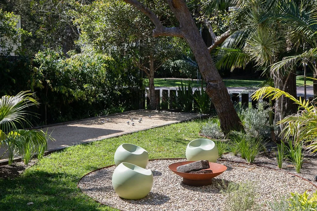 A small outdoor seating area with three modern, white, round chairs surrounding a red fire bowl with a log inside, on a circular gravel patch, amidst lush green trees and plants in a garden.